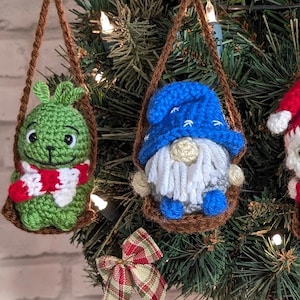 May include: Four crocheted Christmas decorations hanging from a tree. The decorations are a green Grinch-like character, a blue gnome, a red Santa Claus, and a white snowman. Each decoration is sitting in a brown crocheted swing.