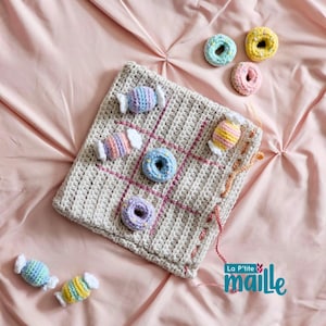 Puede incluir: Un tablero de juego de tres en raya tejido a crochet con rayas rosas y blancas. El tablero está hecho de hilo beige y tiene nueve casillas. Hay caramelos y rosquillas de colores tejidos a crochet en el tablero. El texto "La P'tite maille" es visible en la esquina inferior derecha de la imagen.