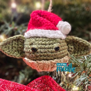 Peut inclure: Ornement crocheté représentant un personnage aux grandes oreilles, portant un bonnet de Père Noël rouge et blanc. L'ornement est vert, beige et rouge, avec des yeux noirs. Il est suspendu à une ficelle, avec un sapin de Noël en arrière-plan.