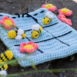 Peut inclure: Un jeu de tic-tac-toe en crochet bleu avec des lignes noires et des abeilles en crochet jaunes et blanches et des fleurs en crochet roses.