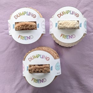 May include: Three round, plush, crocheted dumpling toys in shades of tan and cream. Each toy is wrapped in a white paper band labelled DUMPLING FRIEND with rainbow-coloured text and small cartoon dumpling illustrations on the sides.