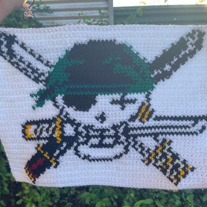 May include: White crochet square featuring a skull with a green bandana and two crossed swords. The swords have yellow and black details. The skull has a black eye patch and is surrounded by a white border.