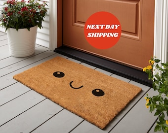 May include: A rectangular doormat made of natural coir fiber. The mat features a smiling face design with black eyes and a curved mouth. The text "NEXT DAY SHIPPING" is in a red circle near the door.
