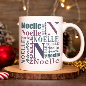 May include: White ceramic mug with a design of the name "Noelle" repeated in various sizes and colors, including purple, teal, and gray. The name is arranged in a circular pattern around a large purple "N".