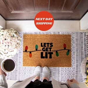 May include: A brown coir doormat with the text "Lets Get Lit" in black lettering. The text is surrounded by a string of colorful Christmas lights.
