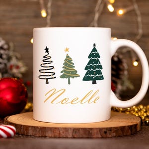 May include: White ceramic mug with a Christmas tree design and the name "Noelle" in gold lettering.