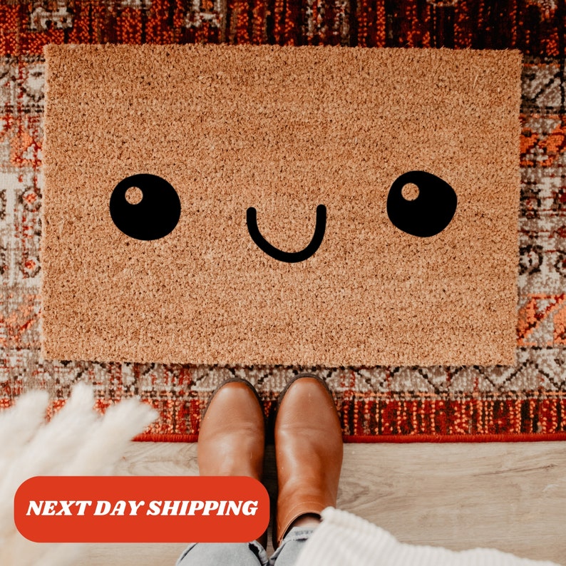 May include: A rectangular doormat made of natural coir fiber features a smiling face design. The face has two large black oval eyes and a curved black mouth. The doormat is placed on a patterned rug, with a person's feet and boots visible.