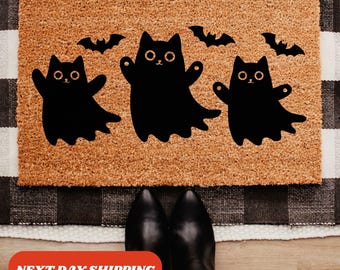 Puede incluir: Una alfombra rectangular con una superficie de fibra de coco marrón presenta tres figuras de gato fantasma negro y tres siluetas de murciélagos. El texto "NEXT DAY SHIPPING" se muestra en una pancarta roja en la parte inferior de la imagen.
