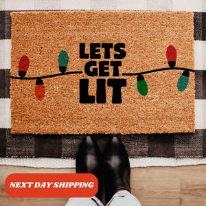 May include: A brown coir doormat with the text "Lets Get Lit" in black lettering. The text is surrounded by a string of colorful Christmas lights.