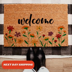 May include: A welcome mat with a brown coir surface and the word "welcome" in black script. The mat is decorated with a row of colorful flowers along the bottom.