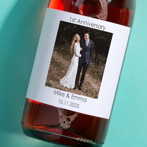 May include: A white label with a photo of a couple in wedding attire, the text "1st Anniversary", "Mike & Emma", and the date "10.11.2025". The label is attached to a bottle of red wine.