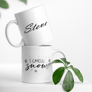 Puede incluir: Dos tazas de cer&aacute;mica blanca. La taza de arriba tiene el nombre "Steve" escrito en cursiva negra. La taza de abajo tiene el texto "I smell snow" escrito en cursiva negra con copos de nieve y estrellas alrededor del texto.