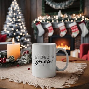 Puede incluir: Taza de caf&eacute; de cer&aacute;mica blanca con texto negro que dice "I smell snow" y dise&ntilde;os de copos de nieve. La taza est&aacute; sobre una mesa de madera con un mantel individual gris.