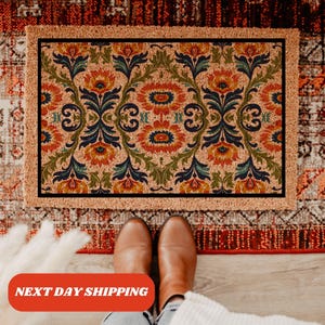 Pu&ograve; includere: Un zerbino rettangolare con sfondo beige e motivo floreale simmetrico arancione, blu e verde. Il tappeto ha un bordo marrone e una cornice interna nera. Il testo "NEXT DAY SHIPPING" &egrave; visibile in basso.