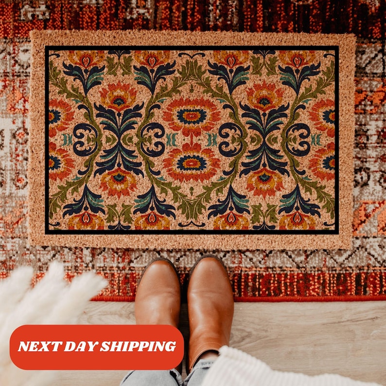 Pu&ograve; includere: Un zerbino rettangolare con sfondo beige e motivo floreale simmetrico arancione, blu e verde. Il tappeto ha un bordo marrone e una cornice interna nera. Il testo "NEXT DAY SHIPPING" &egrave; visibile in basso.