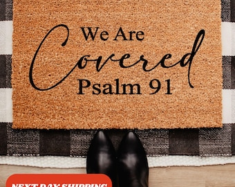 Może przedstawiać: Prostokątna wycieraczka z napisem "We Are Covered Psalm 91" czarnym pismem. Mata jest w jasnobrązowym kolorze i ma teksturowaną powierzchnię. Wycieraczka jest umieszczona na czarno-białym dywanie w kratę.