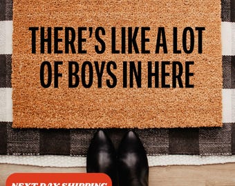 Op de afbeelding: Een rechthoekige deurmat met de zwarte tekst "THERE'S LIKE A LOT OF BOYS IN HERE". De bruine mat ligt op een zwart-wit geruit tapijt. Zwarte laarzen zijn zichtbaar.