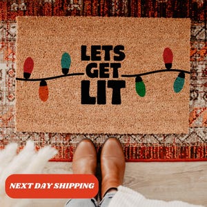 May include: A brown coir doormat with the text "Lets Get Lit" in black lettering. A string of colorful Christmas lights is strung across the top of the doormat.