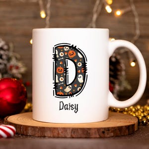 May include: A white ceramic mug featuring a large letter D filled with a Halloween-themed pattern of pumpkins, ghosts, eyeballs, and candy corn. The letter is outlined in black with stitch-like details. The name Daisy is printed below in black.