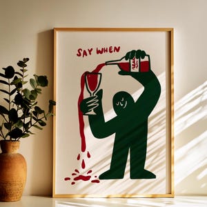 May include: A framed wall art print featuring a dark green cartoon figure pouring red liquid from a bottle into a glass. The text "SAY WHEN" is printed in red above the figure. The red liquid overflows from the glass, creating a spill at the bottom.