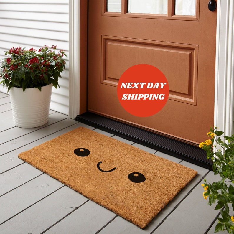 May include: A rectangular doormat made of natural coir fiber. The mat features a smiling face design with black eyes and a curved mouth. The text "NEXT DAY SHIPPING" is in a red circle near the door.