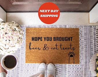 May include: A brown coir doormat with the text "HOPE YOU BROUGHT beer & cat treats" in black lettering. A black paw print is to the right of the text.