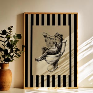 May include: Black and white framed art print featuring a frog sitting on a toilet and reading a newspaper titled "Frog News". The artwork has a beige background and a black and white striped border.