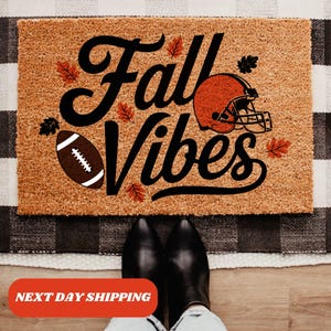 May include: A brown coir doormat with the text "Fall Vibes" in black script. A football and a brown and orange football helmet are on the doormat.