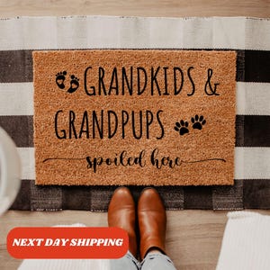 May include: A brown coir doormat with the text "Grandkids & Grandpups Spoiled Here" in black lettering. The text includes a footprint symbol and a paw print symbol.