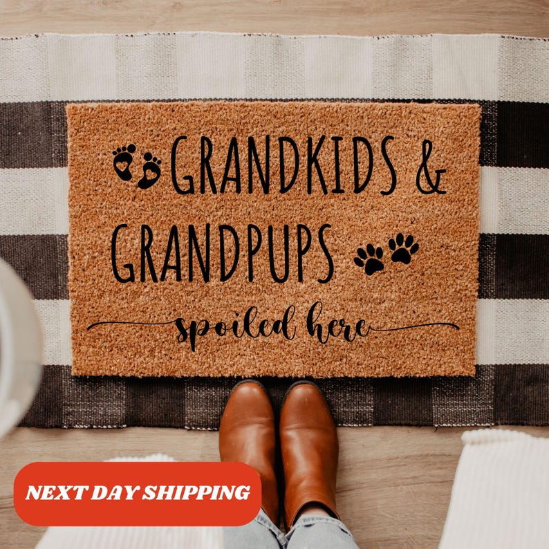 May include: A brown coir doormat with the text "Grandkids & Grandpups Spoiled Here" in black lettering. The text includes a footprint symbol and a paw print symbol.