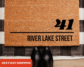 May include: A rectangular doormat made of natural coir fiber, displaying the address "41 River Lake Street" in bold black font. The mat is placed on a black and white checkered rug, with a person's feet in black boots visible.