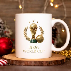 May include: A white ceramic mug featuring a gold-colored graphic of the World Cup trophy, a soccer ball, and laurel branches. Above the trophy are three small stars. The text 2026 WORLD CUP is printed in gold lettering below the graphic.