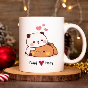 May include: White ceramic mug featuring a cartoon panda and bear design with pink hearts. The mug has the names "Frank" and "Daisy" with a heart symbol. The mug is on a wooden coaster.