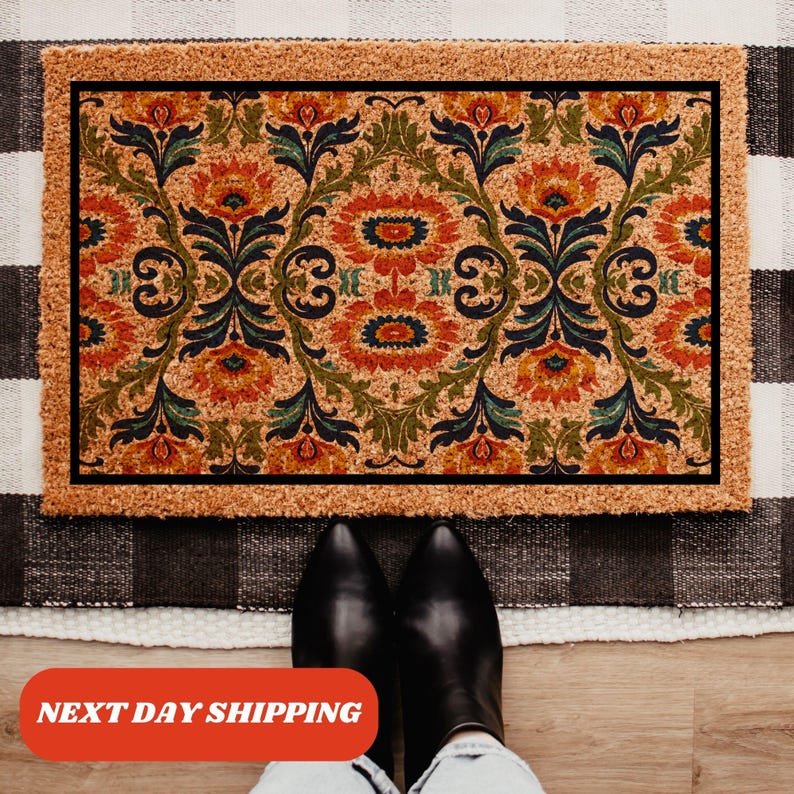 Pu&ograve; includere: Zerbino rettangolare con un motivo floreale nei toni dell'arancione, verde e blu su sfondo beige. Il tappeto ha un bordo nero e una texture naturale in fibra di cocco. Il testo "NEXT DAY SHIPPING" &egrave; visibile.