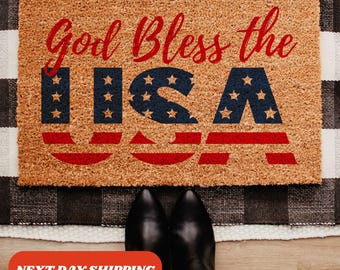 Op de afbeelding: Een bruine kokosvezel deurmat met de tekst "God Bless the USA" in rode en blauwe letters. De letters zijn versierd met rode en blauwe sterren.
