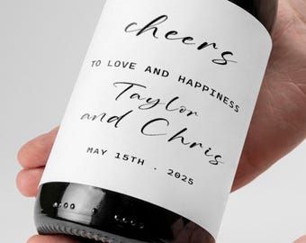 May include: A black wine bottle with a white label. The label reads "Cheers, To Love and Happiness, Taylor and Chris, May 15th, 2025" in elegant black script. The bottle is held by a person.
