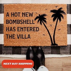 May include: A rectangular tan coir doormat featuring the black text A HOT NEW BOMBSHELL HAS ENTERED THE VILLA. The design includes two black palm tree silhouettes on the right side and several small black bird silhouettes scattered in the sky.