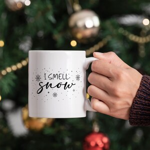 Puede incluir: Taza de cer&aacute;mica blanca con un dise&ntilde;o negro que dice "I smell snow" con copos de nieve y estrellas alrededor del texto. La taza est&aacute; siendo sostenida por una mano frente a un &aacute;rbol de Navidad.