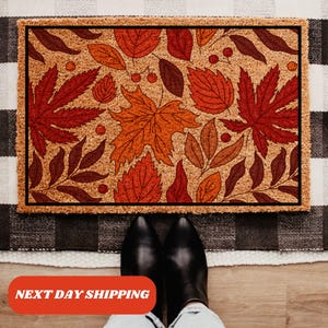 May include: A brown coir doormat with a fall-themed design featuring red, orange, and brown leaves. The leaves are outlined in black and have a stylized look.