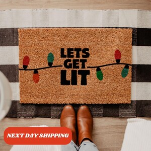 May include: A brown coir doormat with the text "Lets Get Lit" in black lettering. The text is surrounded by a string of colorful Christmas lights.