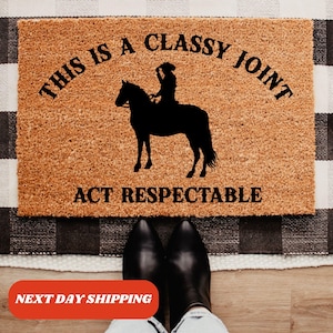 May include: A brown coir doormat with the text "This is a Classy Joint Act Respectable" in black lettering. A silhouette of a person riding a horse is in the center of the mat.