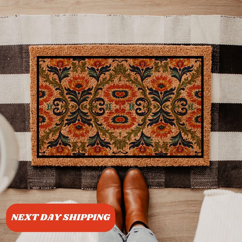 Pu&ograve; includere: Zerbino rettangolare con motivo floreale nei toni dell'arancione, verde e blu su sfondo beige. Ha un bordo nero e una trama in fibra naturale. Il testo "NEXT DAY SHIPPING" &egrave; visibile.