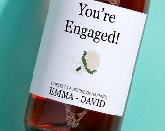 May include: A bottle of rose wine with a white label that says "You're Engaged!" with a white floral bouquet illustration. The label also says "Cheers to a lifetime of happiness Emma - David".