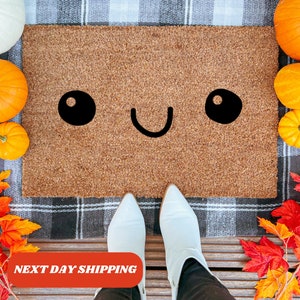 May include: A rectangular coir doormat with a smiling face design. The face has two large black oval eyes and a curved black mouth. The mat is surrounded by pumpkins and fall foliage. The text "NEXT DAY SHIPPING" is at the bottom.