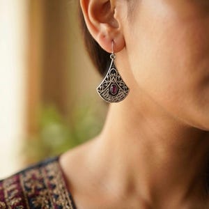May include: A silver-toned dangle earring featuring a fan-shaped design with intricate filigree scrollwork and a central oval-shaped deep red gemstone. The metal surface displays a darkened, oxidized finish within the recessed decorative patterns.