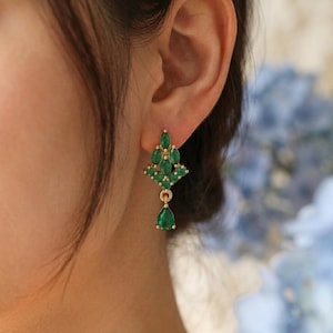 May include: Gold earrings adorned with emerald-green gemstones. The earrings showcase a cluster of smaller stones at the top and a single, teardrop-shaped stone hanging below. The design is elegant and suitable for special occasions.