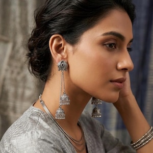 May include: Silver-toned, dangling earrings with a tiered bell design. The earrings are attached to a small, circular stud with a dark center stone. The model is also wearing a layered silver necklace and a silver bracelet.