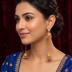 May include: Gold-toned, dangling earrings with a bell-shaped design. The earrings feature intricate detailing and a long chain that connects the bell to a smaller stud. The earrings are worn with a blue top with gold embroidery.