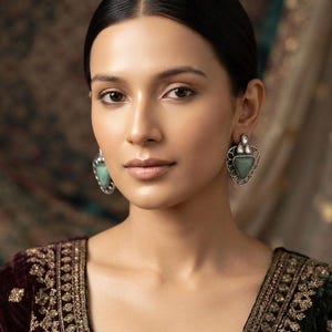 May include: A pair of silver-toned earrings featuring a triangular green stone set within an intricate filigree frame, accented by a small clear teardrop-shaped stone at the top, worn with a dark velvet garment featuring gold embroidery.
