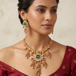 May include: A gold-toned necklace and matching earring set featuring intricate metalwork, circular green enamel centers, and small dangling charms. The necklace includes clear stones and red accents, while the earrings mirror the design.
